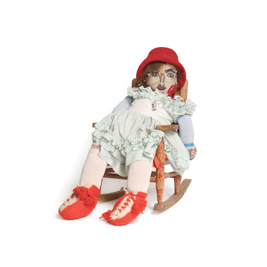 A cloth doll with beige skin wearing a red hat, broken glasses, and a ruffled light green dress sits in a wooden rocking chair.