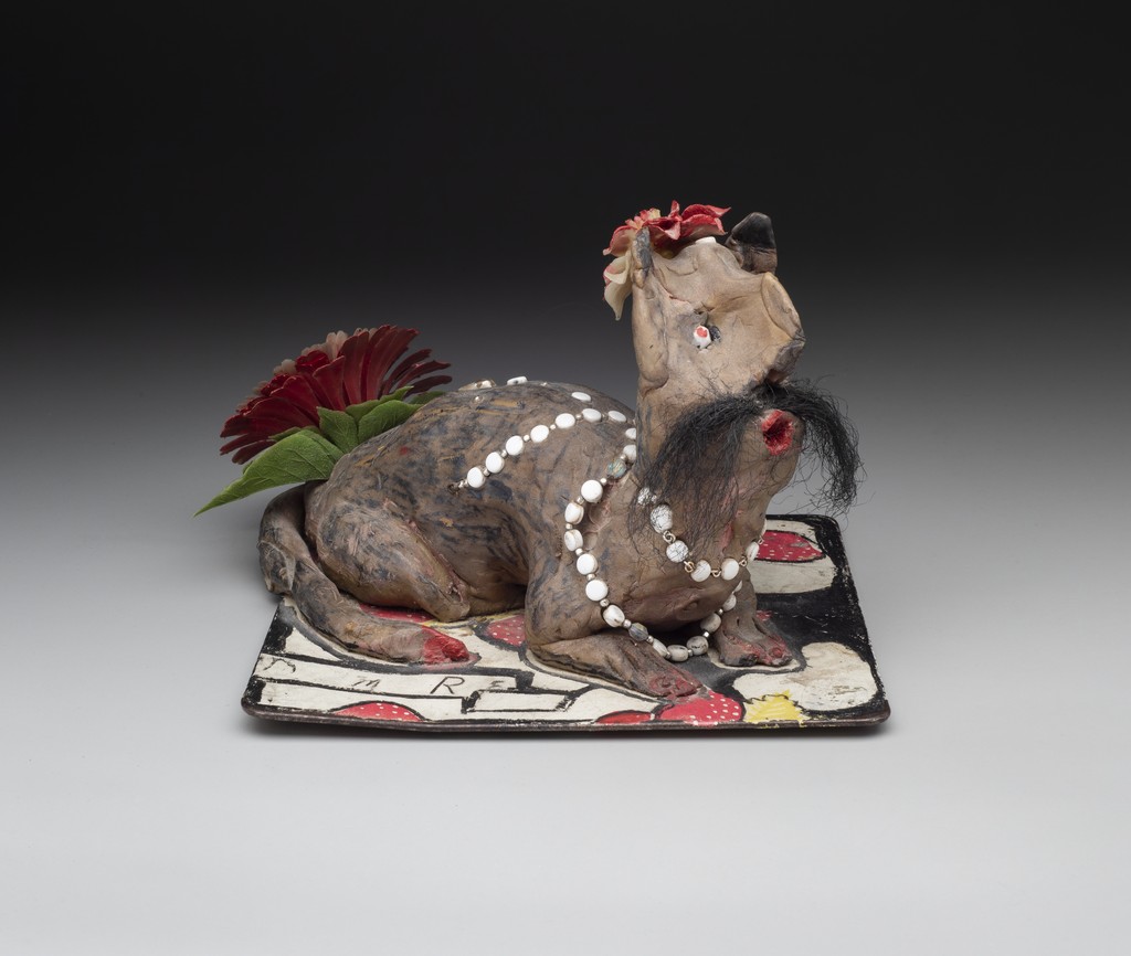 Light purple sculpture of cat-like body and amorphous face with red mouth in O-shape, long mustache of hair, and plastic red flowers at crown of head and back of body. It wears a necklace of white beads and crouches on a flat trivet.