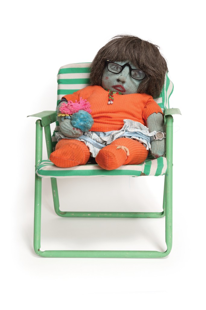 A cloth doll with green skin and shoulder-length brown hair wearing glasses, an orange shirt, and a watch sits in a wooden rocking chair.